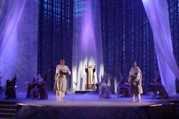 white long scarves with purple wash lights on them and on a stage with low fog cover with people sitting in with a performer in robes with hands up in the air and a white spotlight on them, two performers in robes holding props, Gabriel's Christmas Story, Calvary Orlando, Winter Park, Florida, USA