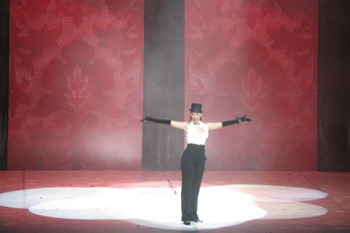 singer in a tophat with long black gloves on and her arms reached out with a white sleevless tuxedo without a coat and several white spotlights on her, behind the performer two large damask floral backdrop panels with burgundy hues on the stage in the play Glamur, Sexo, Divas y Otras Mentiras - Teatro Nacional, Dominican Republic